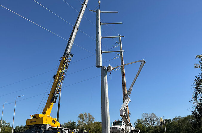 A yellow crane sets a metal electrical pole