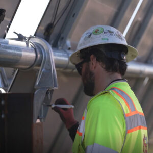 A man in a hard hat and a high-vis vest writes on the back of a solar panel