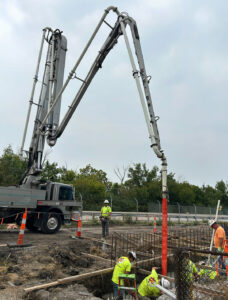 A concrete truck with a large arm pours concrete into the ground as construction workers distribute the concrete